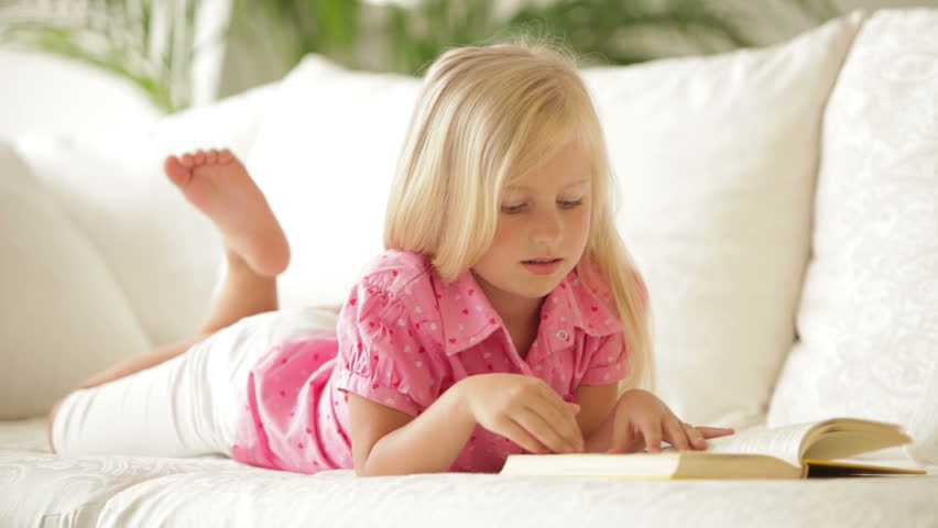Cute little girl lying on sofa reading book and smiling