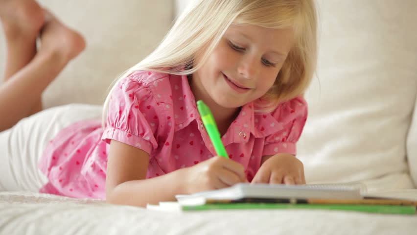 Cheerful little girl lying on sofa writing in notebook and smiling