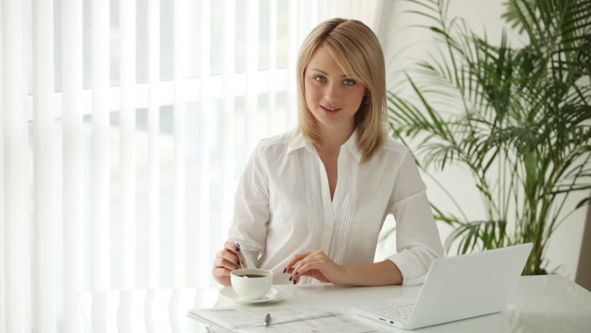Charming young woman sitting at table with laptop stirring coffee and smiling