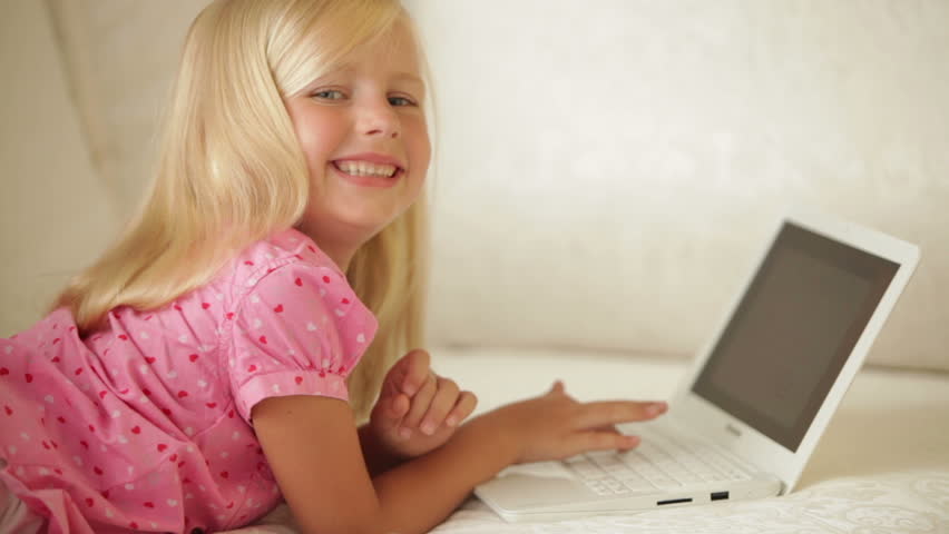 Beautiful little girl lying on sofa using laptop and smiling at camera