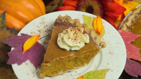 Slice of pumpkin pie, topped with whipped cream, displayed, rotating, above festive holiday background of whole pie, fall leaves, red sunflower, squash.  Stock Video
