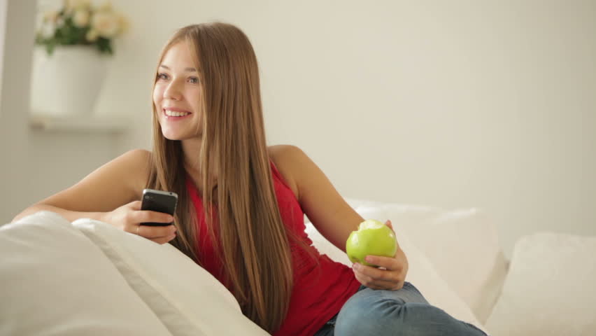 Cute girl sitting on sofa eating apple and using cellphone
