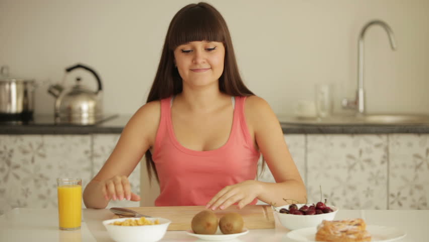 Cheerful girl sitting at kitchen table and peeling kiwi fruit