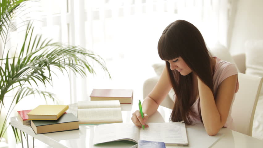 Cheerful girl studying at table with books writing in notebook and smiling at