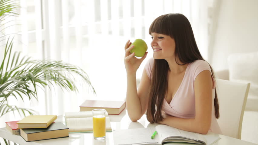 Charming student girl studying at table holding apple and smiling