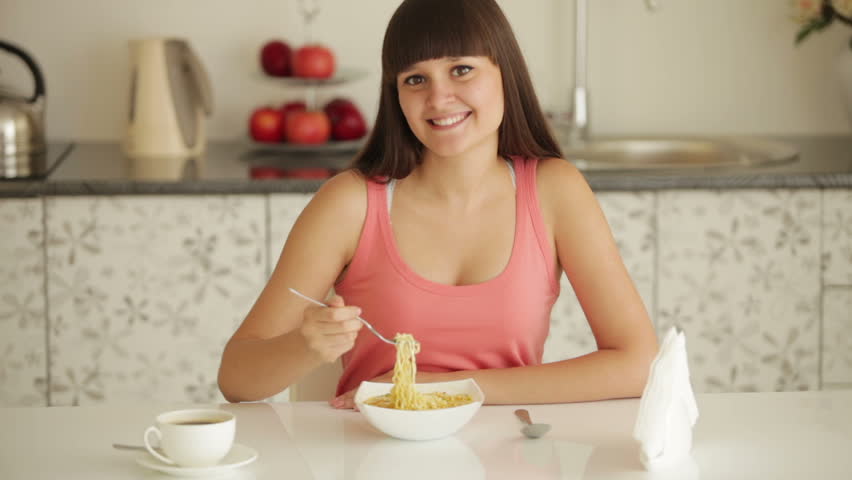 Cheerful girl sitting at kitchen table and eating noodle