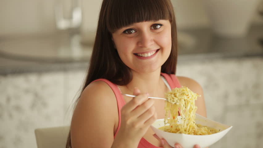 Cheerful girl at kitchen eating noodle and smiling