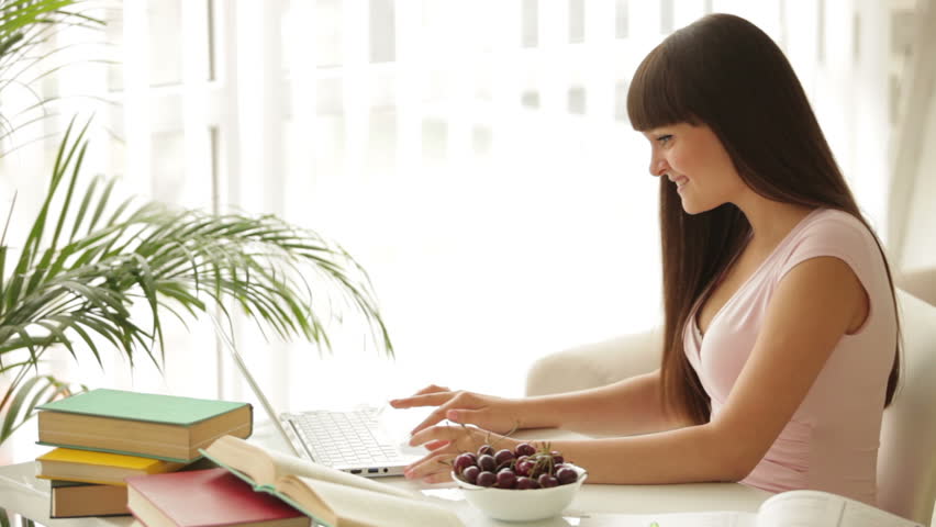 Charming student girl sitting at table using laptop and eating cherries