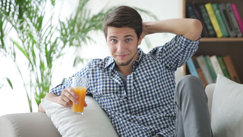 Charming guy sitting on sofa holding glass of juice and smiling
