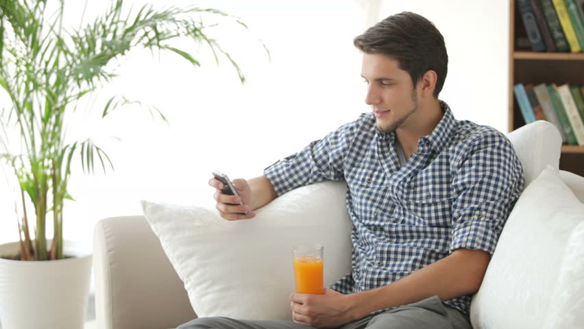 Cheerful guy sitting on sofa using mobile phone and drinking juice