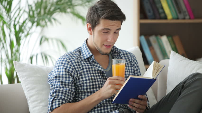 Charming guy sitting on sofa reading book and drinking juice