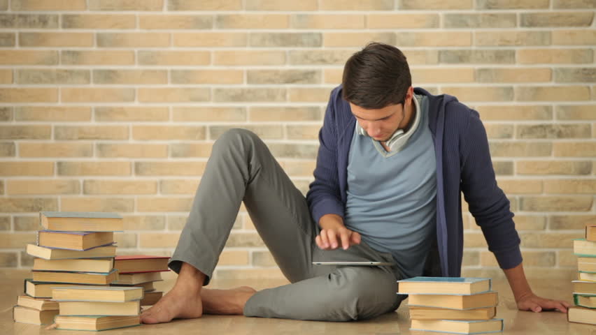 Cheerful guy sitting on floor with stack of books and using touchpad