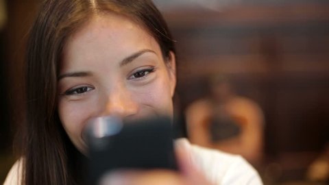Girl using smartphone in cafe drinking coffee laughing in cafe. Beautiful multicultural young female professional having casual conversation on mobile phone. Mixed race Asian Caucasian model.
