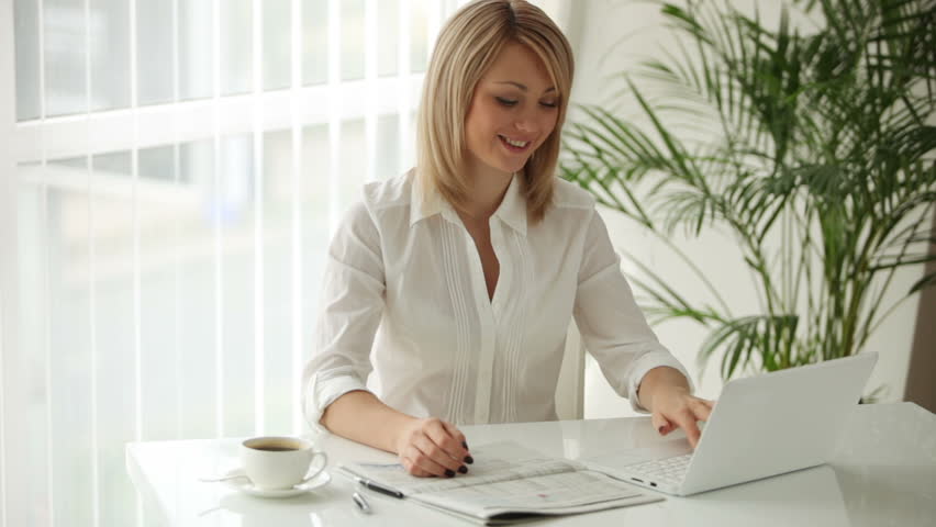 Young businesswoman sitting at table with newspaper and using laptop