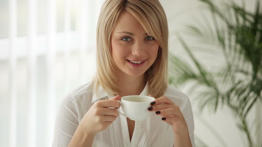 Pretty young woman sitting at table drinking coffee and smiling at camera