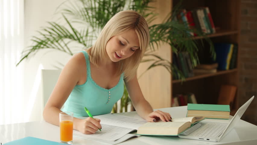 Cheerful girl sitting at table studying with books and laptop and smiling at