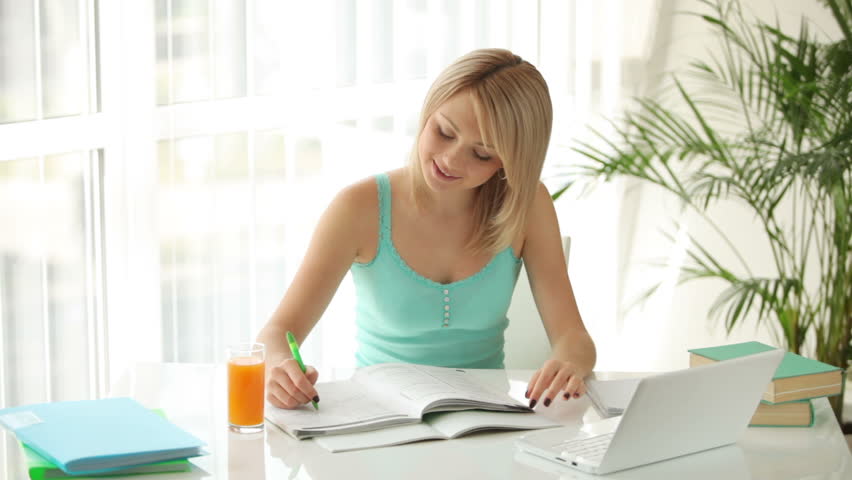 Cute girl sitting at table writing in notebook drinking juice and smiling at