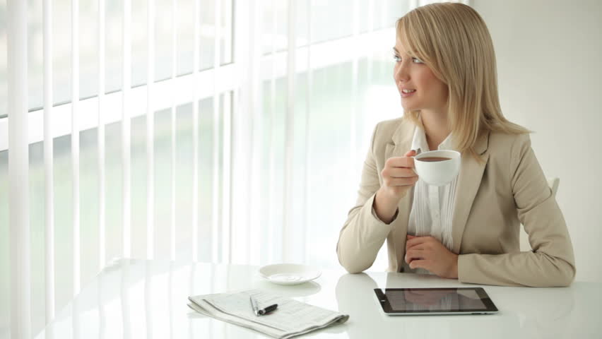 Cheerful young woman sitting at office table drinking coffee and smiling