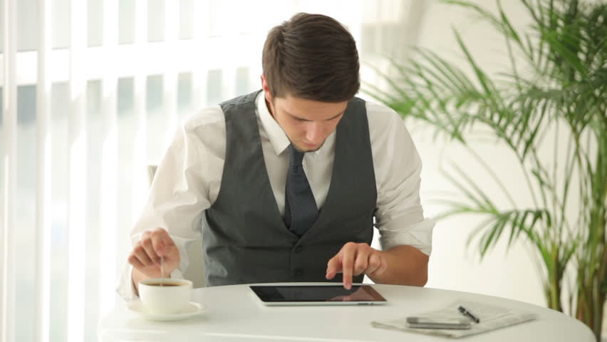 Handsome man sitting at table stirring coffee and using touchpad