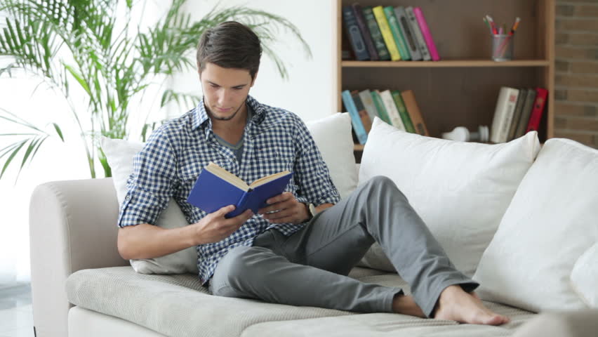 Handsome young man sitting on sofa with book and smiling