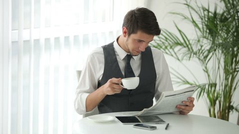 Good-looking man sitting at table drinking coffee and reading newspaper