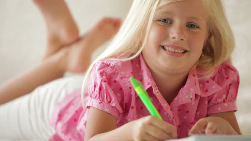 Pretty little girl lying on sofa writing in notebook and smiling at camera