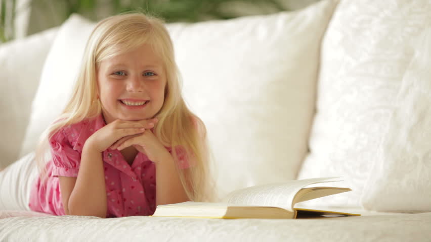 Happy little girl lying on sofa with book and smiling at camera