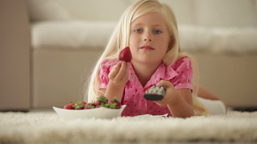 Pretty little girl lying on floor eating strawberries and holding remote control