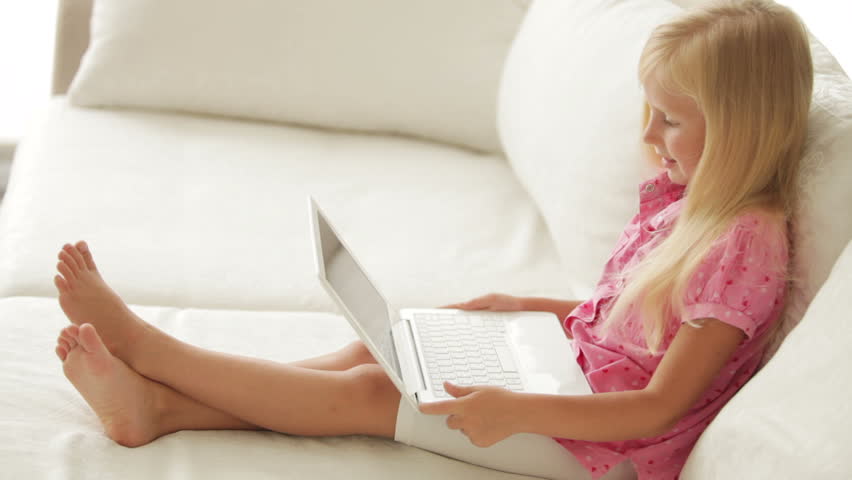 Pretty little girl sitting on sofa using laptop and smiling at camera