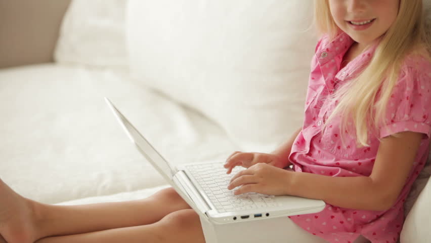 Funny little girl sitting on sofa using laptop and smiling