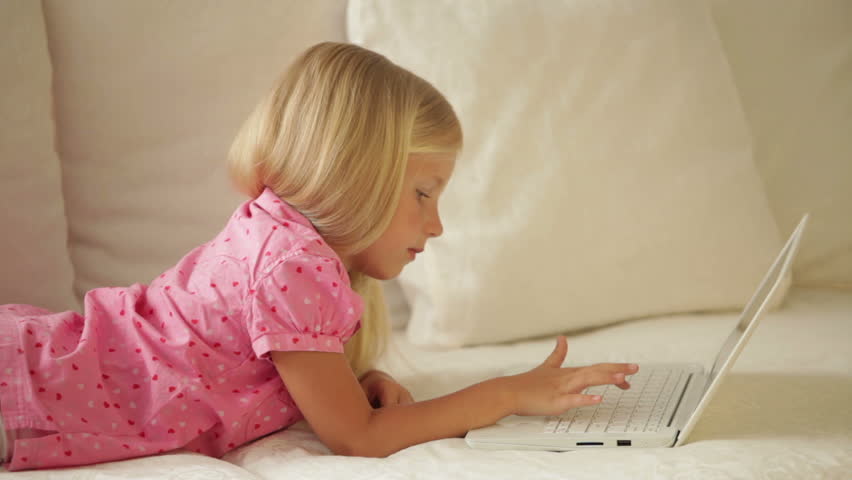 Pretty little girl relaxing on sofa using laptop and smiling at camera