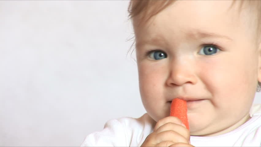 Baby sits at a table and eats a carrot. Isolated on the white