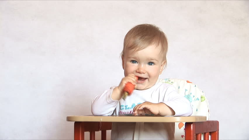 Baby sits at a table and eats a carrot. Isolated on the white
