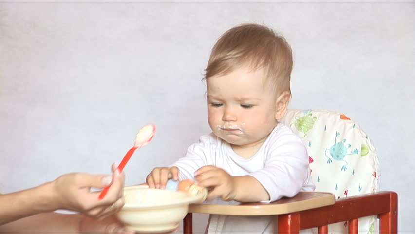 Baby sits at a children's little table. Mum feeds the child with porridge.