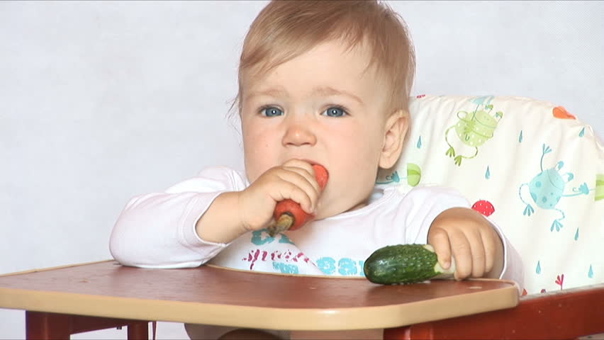 Baby sits at a table and eats a carrot and cucumber. Isolated on the white