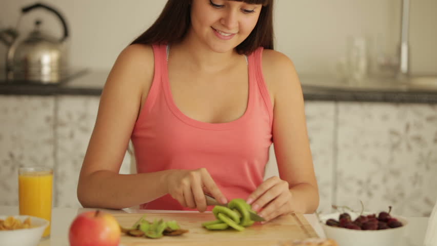 Attractive girl sitting at kitchen table and slicing kiwi