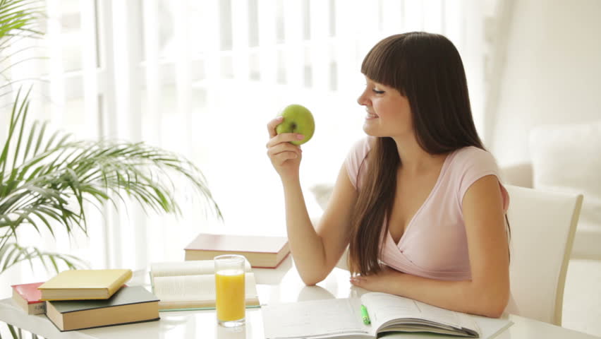 Beautiful girl sitting at table writing in notebook eating apple and smiling