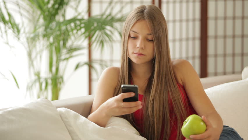 Charming girl sitting on sofa using cellphone and eating apple
