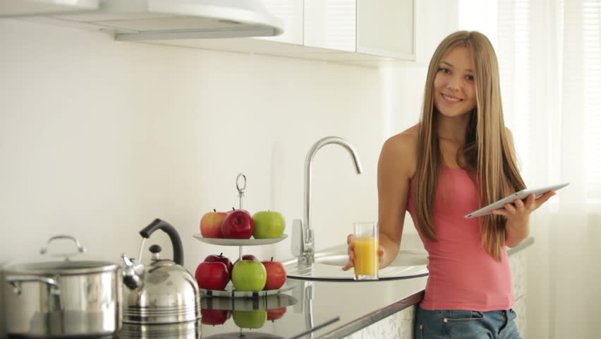 Cheerful girl standing in kitchen drinking juice and using touchpad