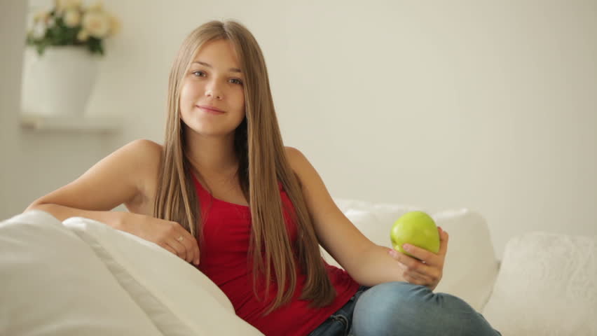 Charming girl sitting on sofa eating apple and smiling at camera