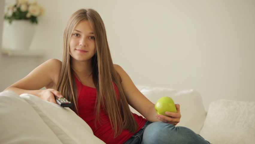 Cute girl on sofa eating apple and using remote control
