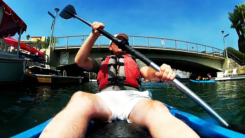 A mature or middle aged man paddles a sea kayak, is kayaking under a bridge on