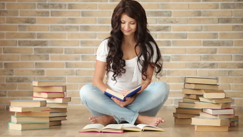 Cute girl sitting on floor with pile of books holding one and smiling