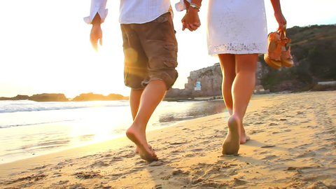 An older couple holds hands and walks down the beach at sunset getting their feet wet