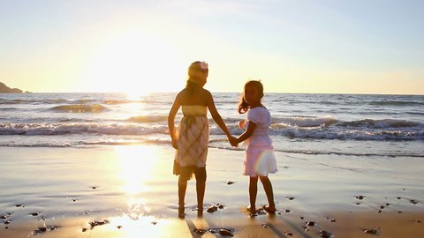 Two young girls hold hands and play with each other while they walk through the water at the beach at dusk