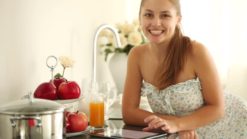 Cute girl standing in kitchen using touchpad looking at camera and smiling.