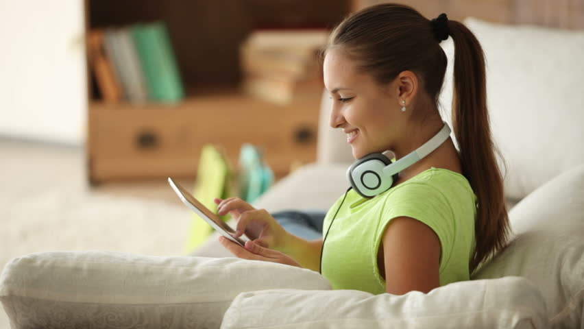 Pretty girl in headphones relaxing on sofa using touchpad and smiling at camera.