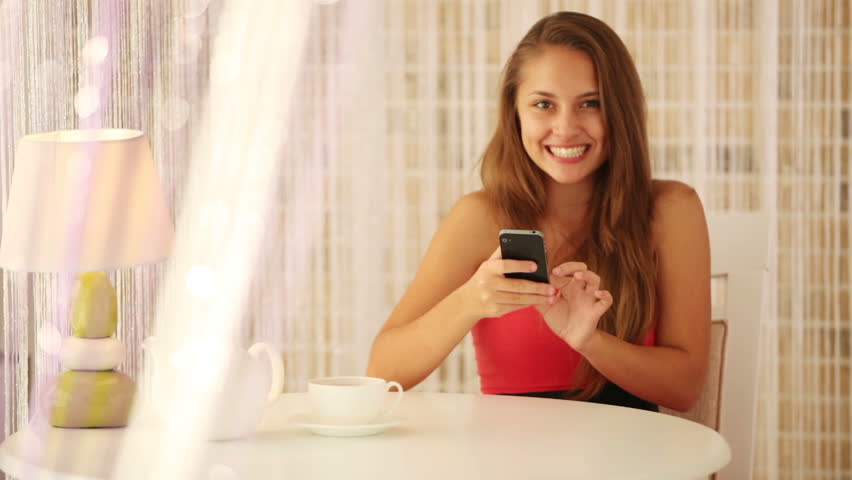 Pretty girl sitting at table at cafe using mobile phone and smiling at camera.