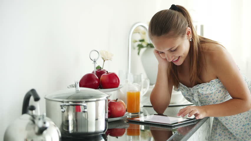 Beautiful girl standing in kitchen using touchpad looking at camera and smiling.