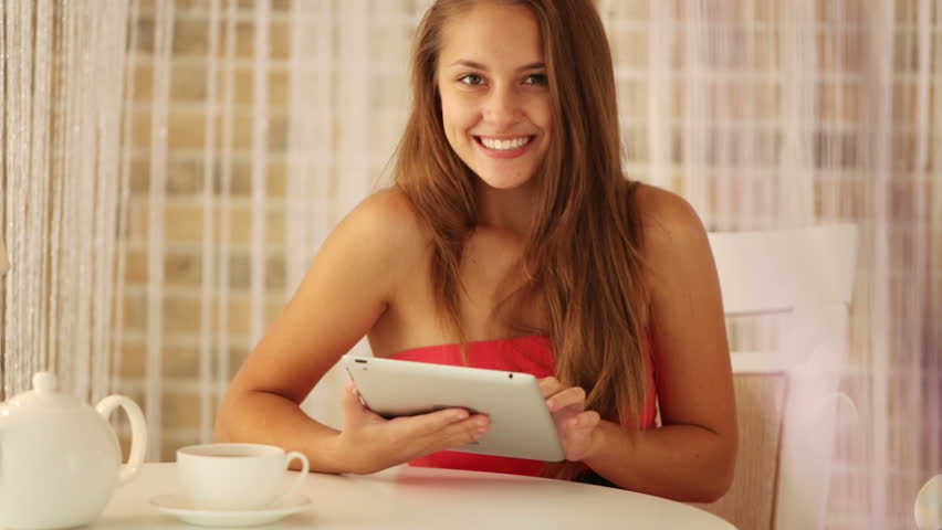 Charming girl sitting at table at cafe using touchpad looking at camera and